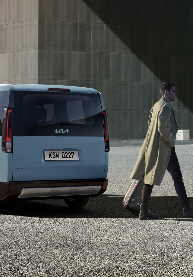 Dreiviertel-Rückansicht des Kia PV5 Passenger, geparkt in der Nähe eines Gebäudes, während ein Mann vorbeigeht.